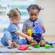 Two young toddlers sit on the carpet in a home daycare as they play together with toy animals and blocks.