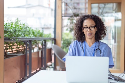 A Black, female doctor wearing scrubs is conducting a tele-health visit with a patient.