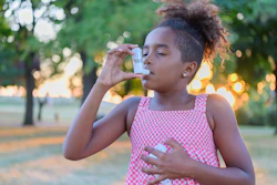 A Black elementary age girl uses her inhaler outdoors.