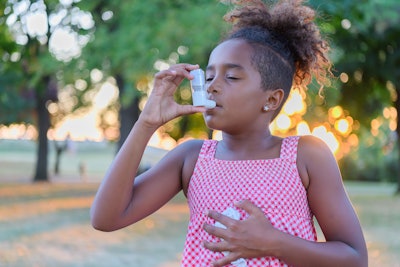 A Black elementary age girl uses her inhaler outdoors.