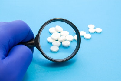 A gloved hand is holding a magnifying glass over some white pills on a counter.