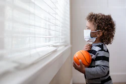A young, Black boy is wearing a mask and standing inside his house looking out the window and holding a ball.