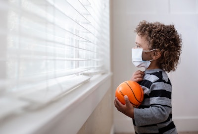 A young, Black boy is wearing a mask and standing inside his house looking out the window and holding a ball.