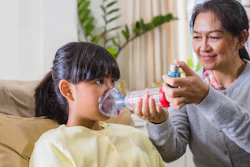 An Asian woman is administering asthma medication through a spacer to an elementary-age girl.