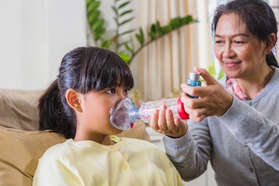An Asian woman is administering asthma medication through a spacer to an elementary-age girl.