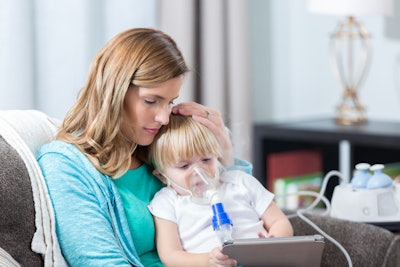 Young girl with cystic fibrosis sits on mom's lap while receiving breathing treatment.