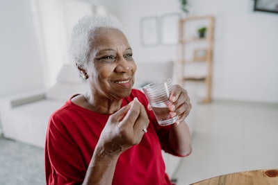 An edlerly, Black woman is taking a pill with a glass of water.