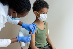 A young Black boy wearing a surgical mask is receiving a shot in the arm by a Black healthcare worker also wearing a mask.