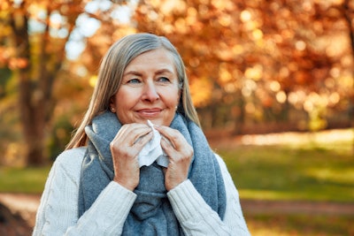 This picture shows a middle-aged, white woman withy grey streaks in her hair smiling outdoors while holding a tissue.