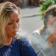 This is a photo of two young, women sitting next together. One is exhaling cigarette smoke and the other is coughing from exposure to second-hand smoke.