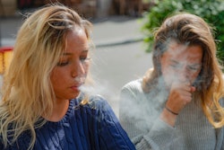 This is a photo of two young, women sitting next together. One is exhaling cigarette smoke and the other is coughing from exposure to second-hand smoke.