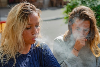 This is a photo of two young, women sitting next together. One is exhaling cigarette smoke and the other is coughing from exposure to second-hand smoke.