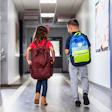 This is a photo of two, multiracial, elementary children walking through the school hallway with their backpacks.
