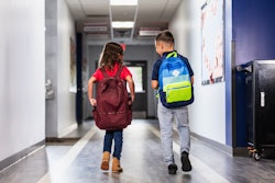 This is a photo of two, multiracial, elementary children walking through the school hallway with their backpacks.