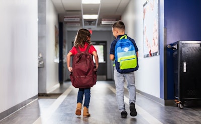 This is a photo of two, multiracial, elementary children walking through the school hallway with their backpacks.