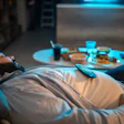 This is a photo of an overweight man asleep on the couch with an array of unhealthy food on a nearby table.