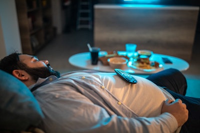 This is a photo of an overweight man asleep on the couch with an array of unhealthy food on a nearby table.