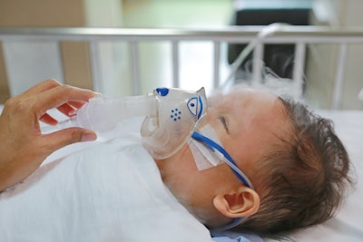 Sick baby boy rest on patients bed and has inhalation therapy by the mask of inhaler.