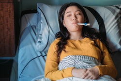 This is a photo of an Asian woman laying in bed sick and taking her temperature with a thermometer.