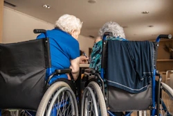 This is a photo of of the back of two, elderly women in wheelchairs in a senior living setting.