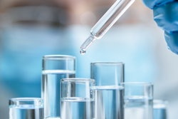 Photo of a gloved hand putting drops of unknown liquid into test tubes with a masked medical person blurred in the background.
