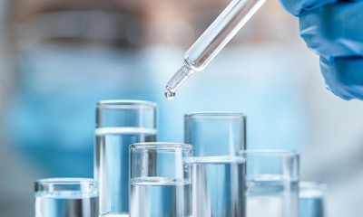 Photo of a gloved hand putting drops of unknown liquid into test tubes with a masked medical person blurred in the background.