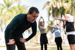This is a photo of a man bent over and struggling to catch his breath while people behind him are exercising,.