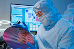 This is a photo of an Asian male technician in sterile coverall holds wafer that reflects many different colors with gloves and check it at semiconductor manufacturing plant.