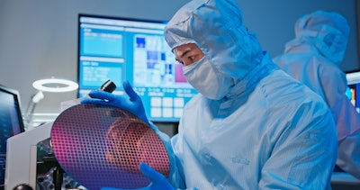 This is a photo of an Asian male technician in sterile coverall holds wafer that reflects many different colors with gloves and check it at semiconductor manufacturing plant.