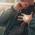 Man sitting on couch holding chest with concerned expression, suggesting health concern or discomfort