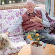 Elderly man relaxing on a purple floral sofa with a small dog beside him and potted flowers nearby