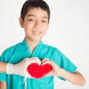 Young boy in teal medical scrubs smiling while forming a red heart shape with his hands