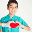 Young boy in teal medical scrubs smiling while forming a red heart shape with his hands
