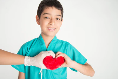 Young boy in teal medical scrubs smiling while forming a red heart shape with his hands