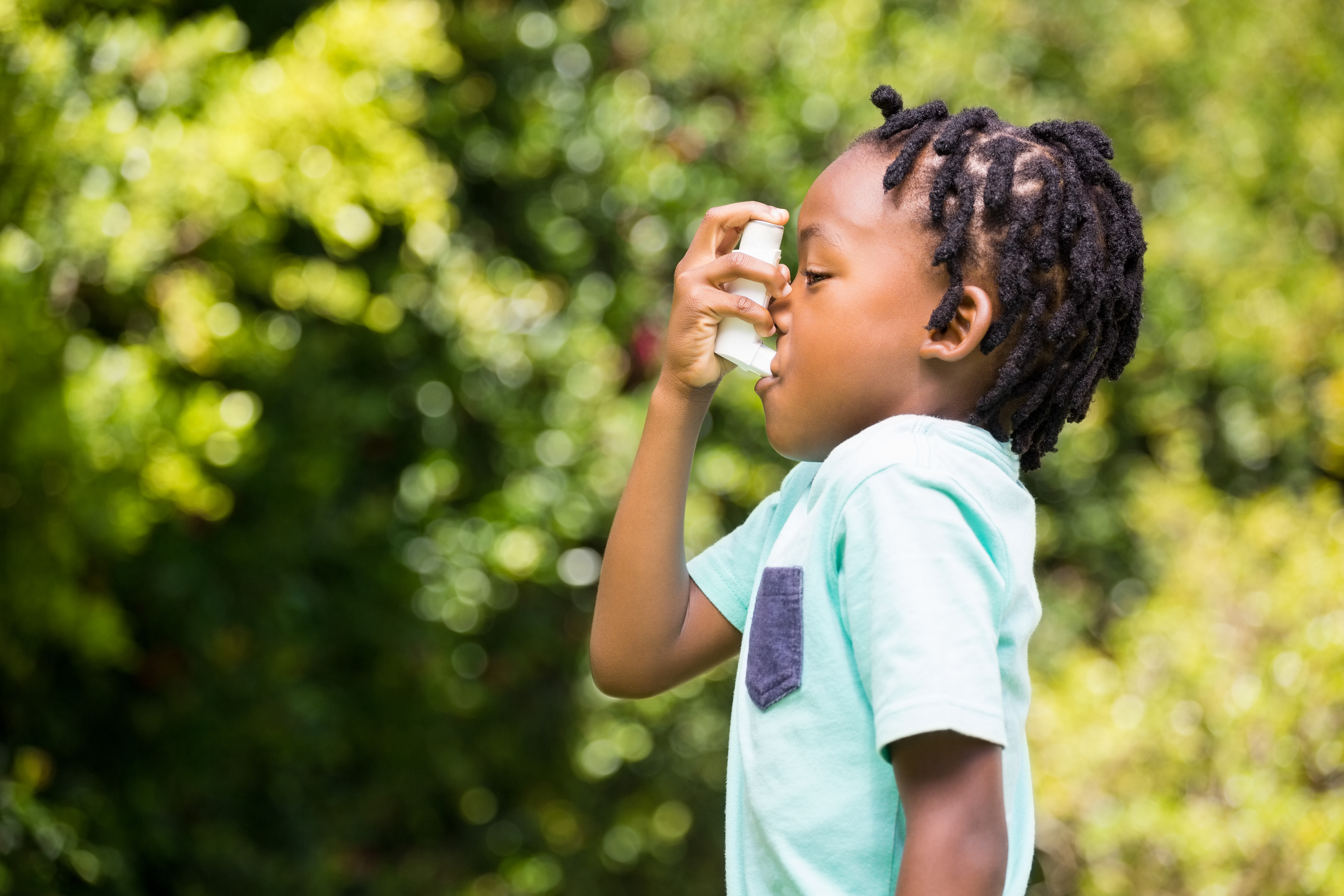 Child using asthma inhaler outdoors with green foliage background