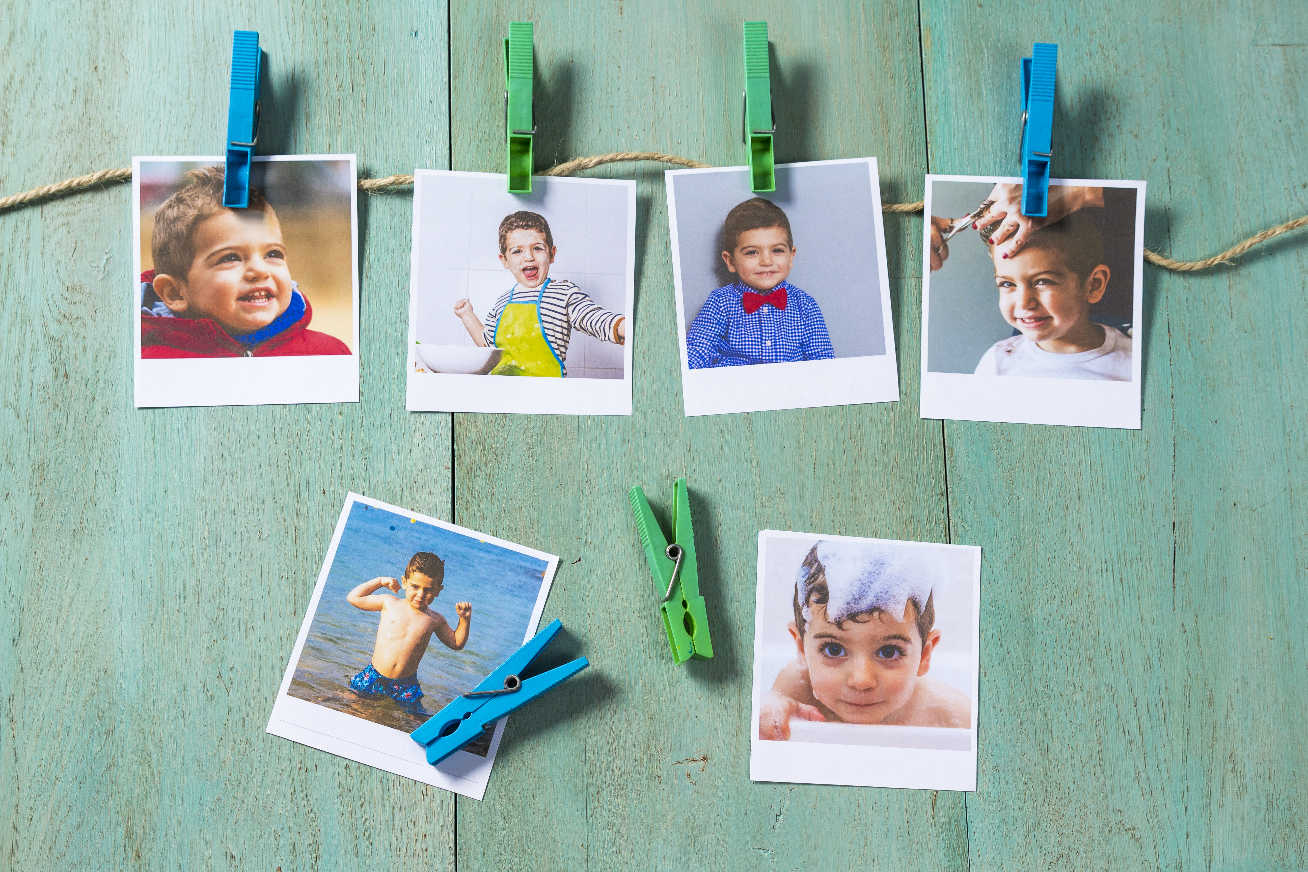 Six printed photos of smiling children clipped to twine with colorful clothespins on a turquoise wooden background