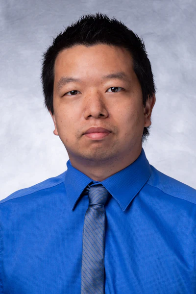 Professional headshot of an Asian man wearing a bright blue dress shirt and patterned tie against a neutral gray background