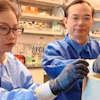 Two researchers in blue lab coats examining samples in a laboratory setting with shelves of scientific equipment and supplies