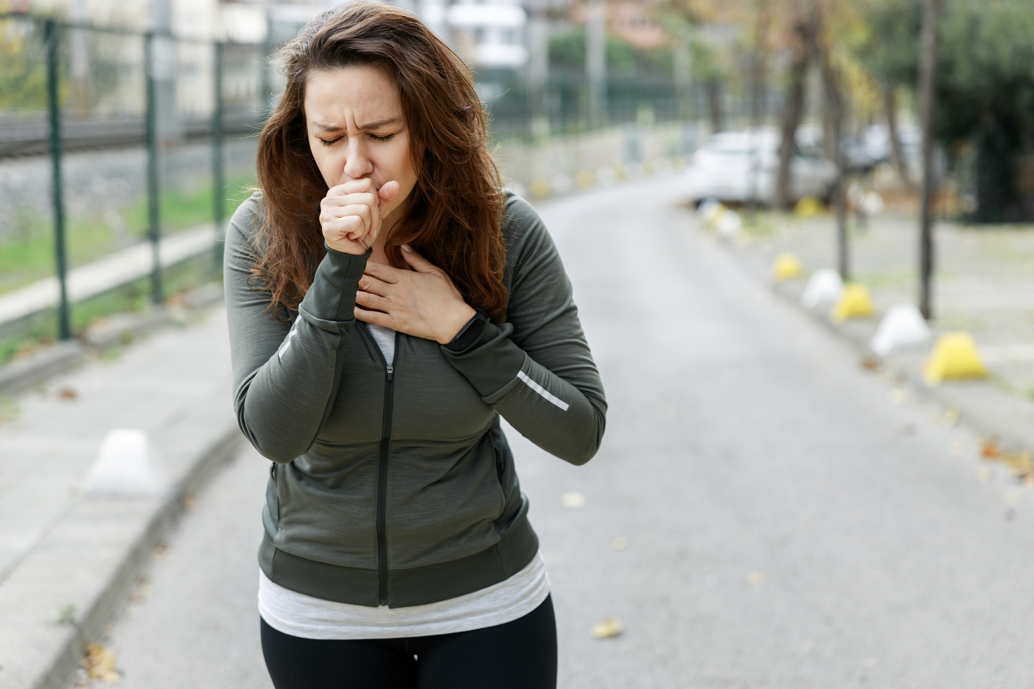 Woman jogging on outdoor path wearing olive green jacket, experiencing chest discomfort while running